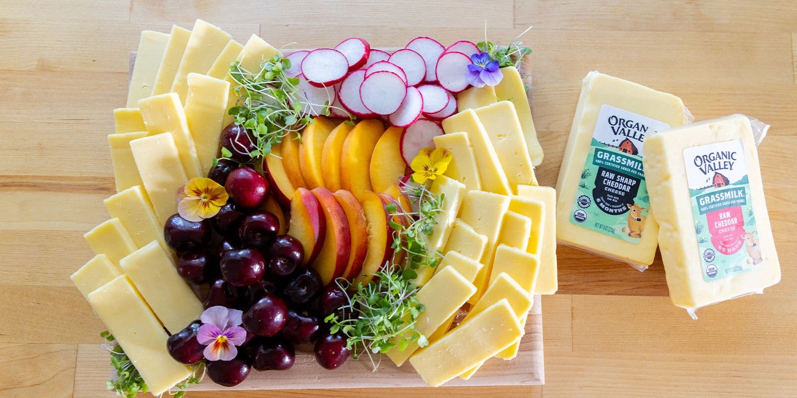 Grassmilk raw: Organic Grassmilk cheeses on a cutting board with peaches, sprouts, cherries, flowers and radishes.