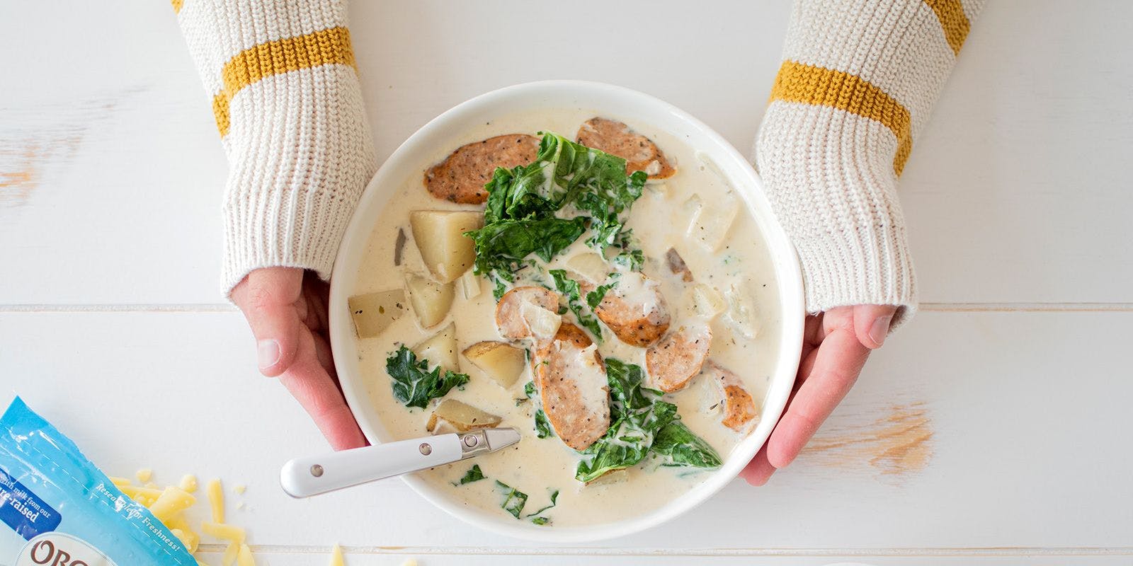 A woman wearing a cozy sweater holds a bowl of sausage potato kale soup.