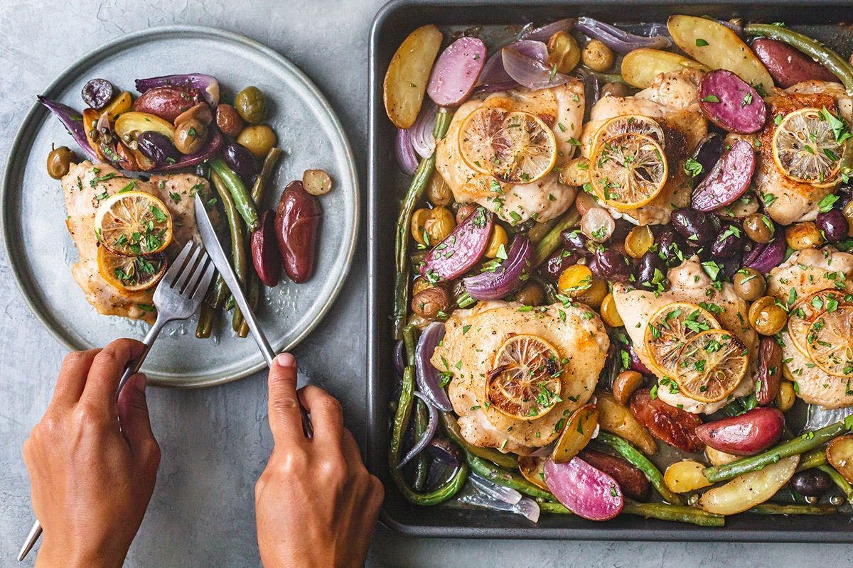 Pan of Chicken Dinner with potatoes and green beans next to a person cutting the chicken on a plate. 