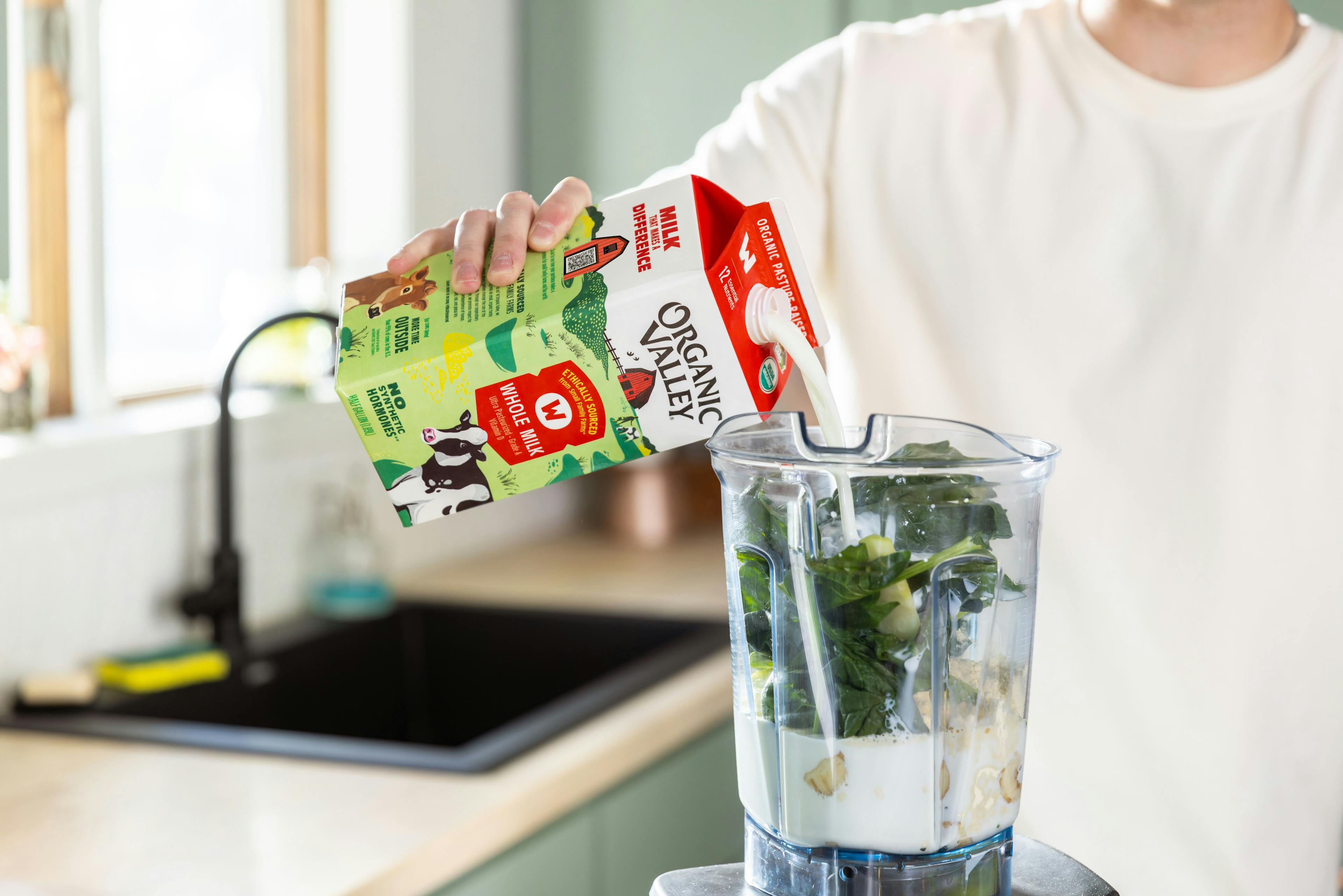 A man pours organic milk in a blender that has greens and other veggies and fruit.