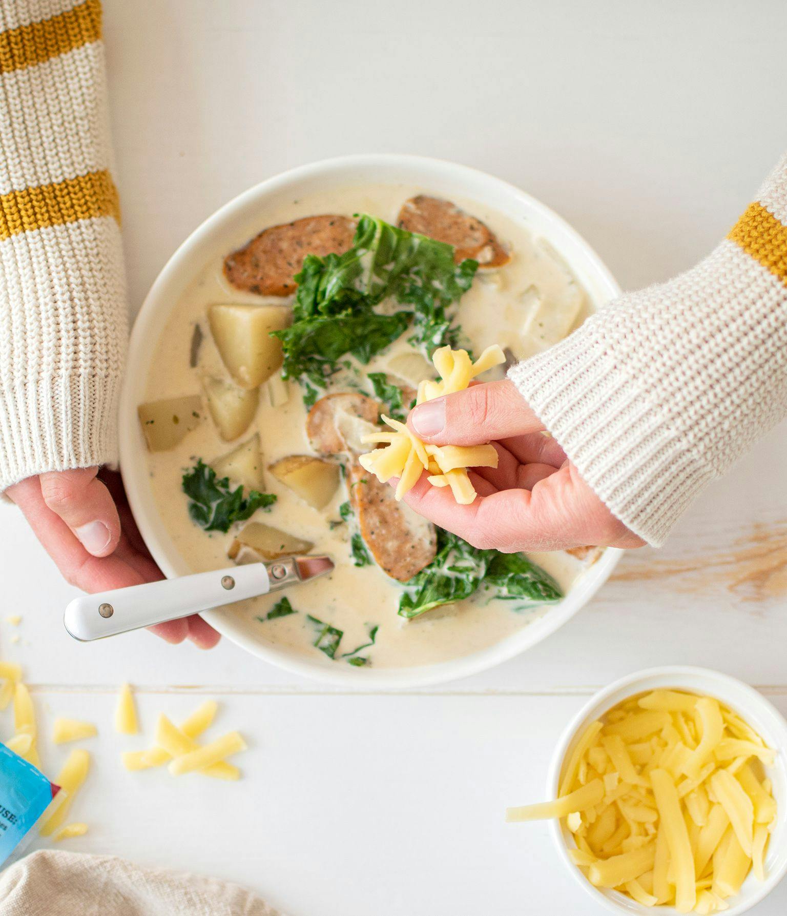 A woman putting Organic Valley cheese into a bowl of Creamy Sausage, Potato, and Kale Soup