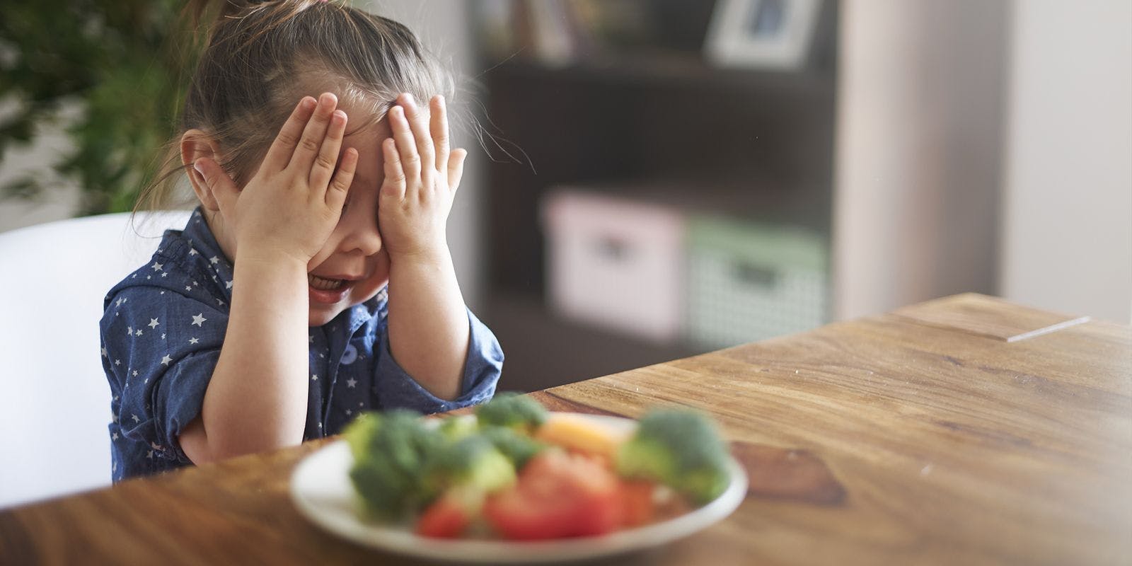 Young girl covers her eyes sitting in front of a plate of food.