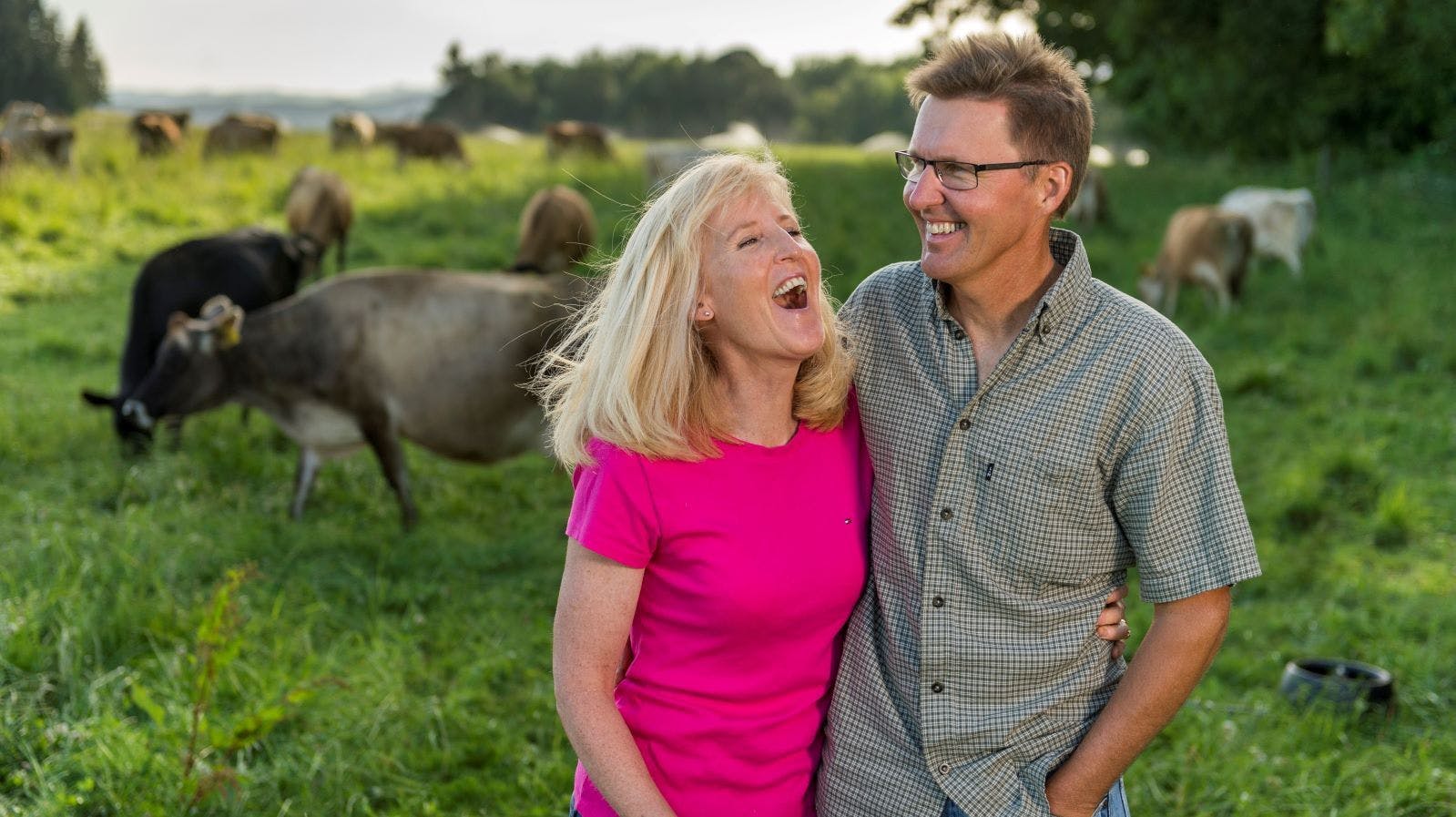 Juli and Jon Bansen stand in a field as cows walk by.