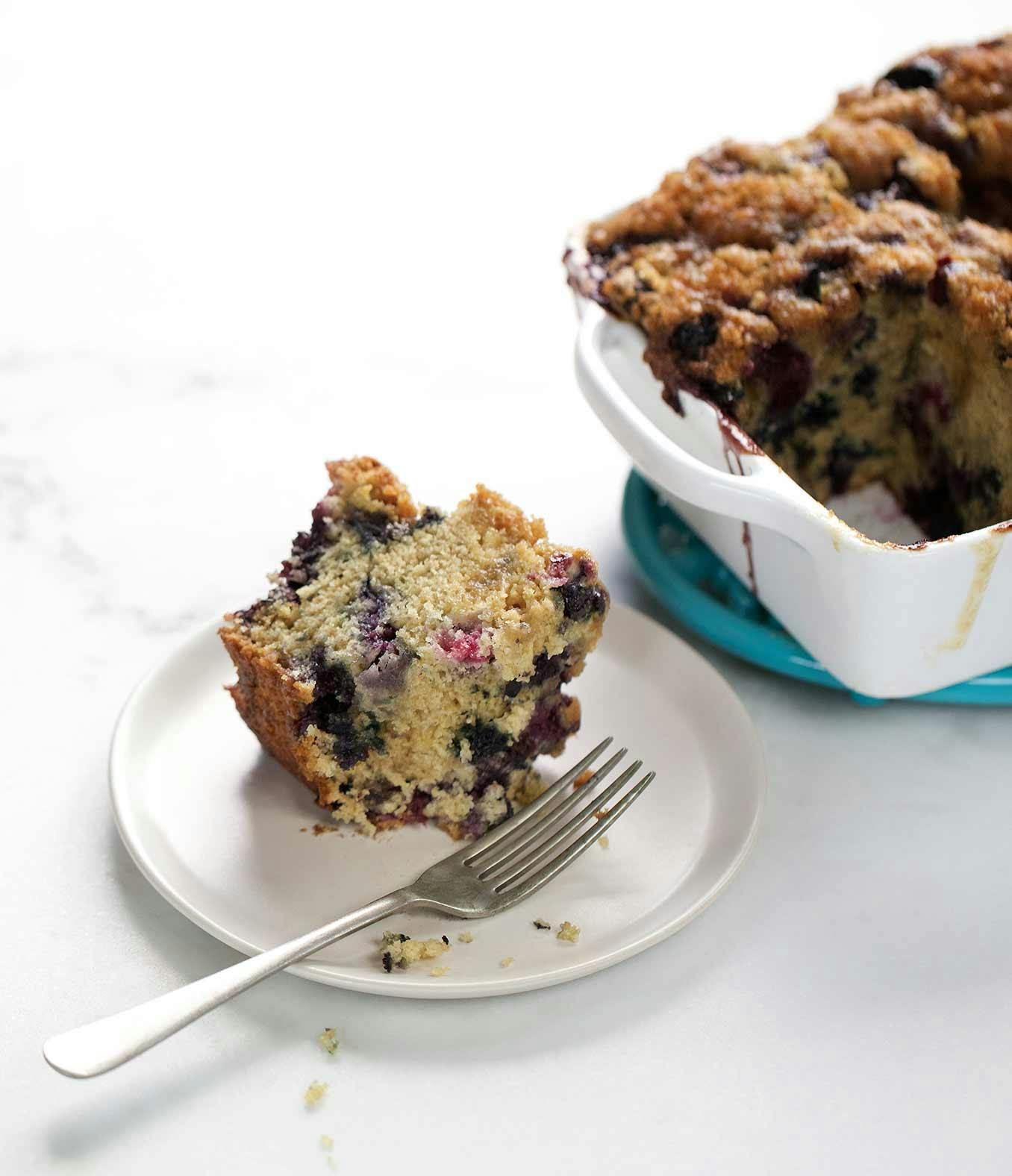 A piece of cream-glazed blueberry coffee cake on a small plate with a fork and the whole cake in a dish on the side.