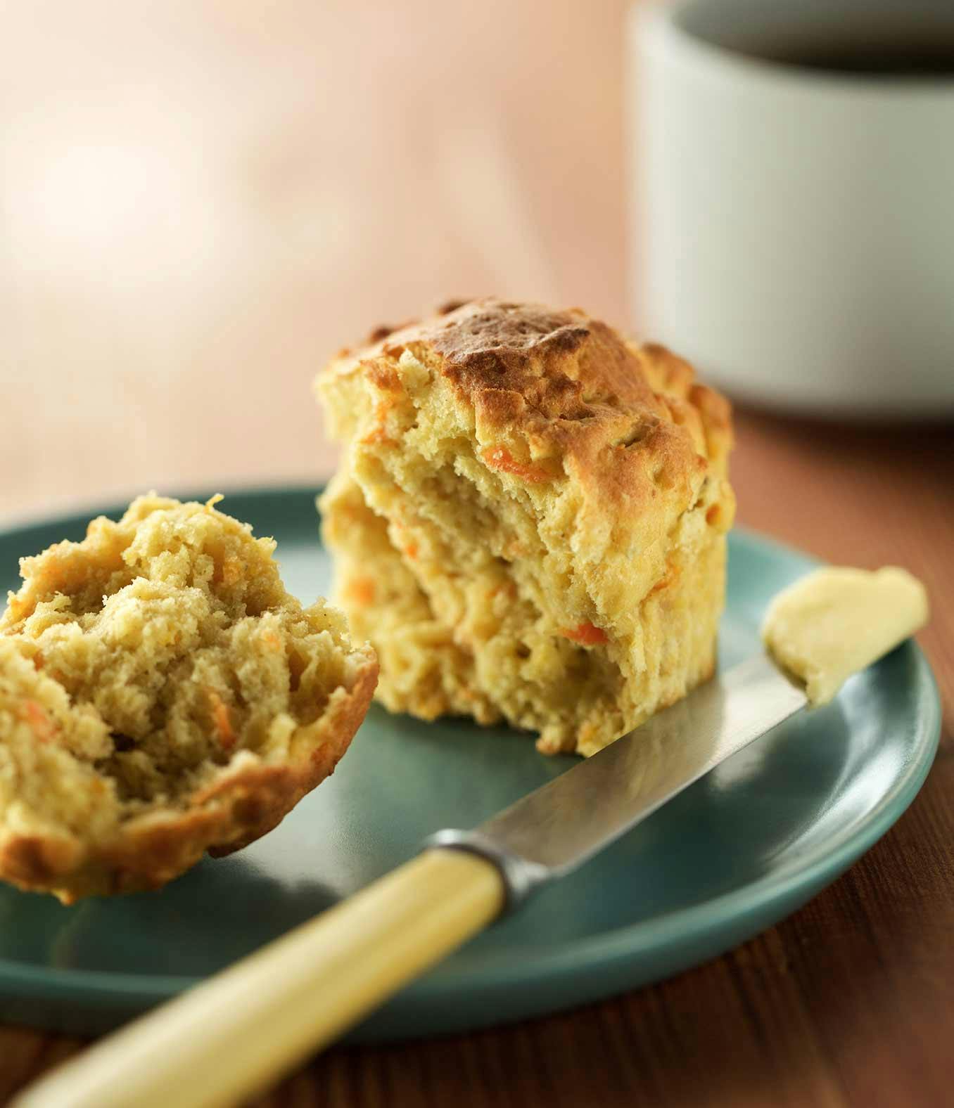 A gluten-free carrot organic muffin cut in half on a plate next to a knife with butter on it.