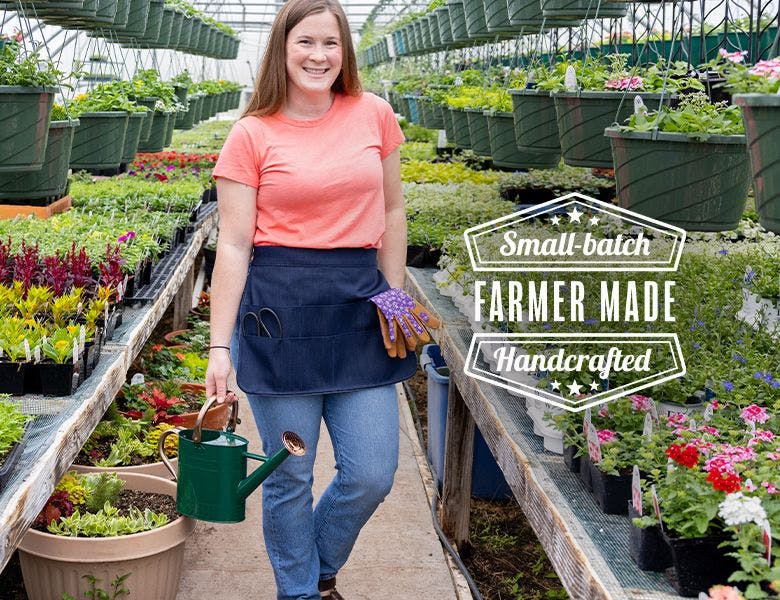 Woman standing in a greenhouse wearing a farmer-made apron and holding a watering can. 